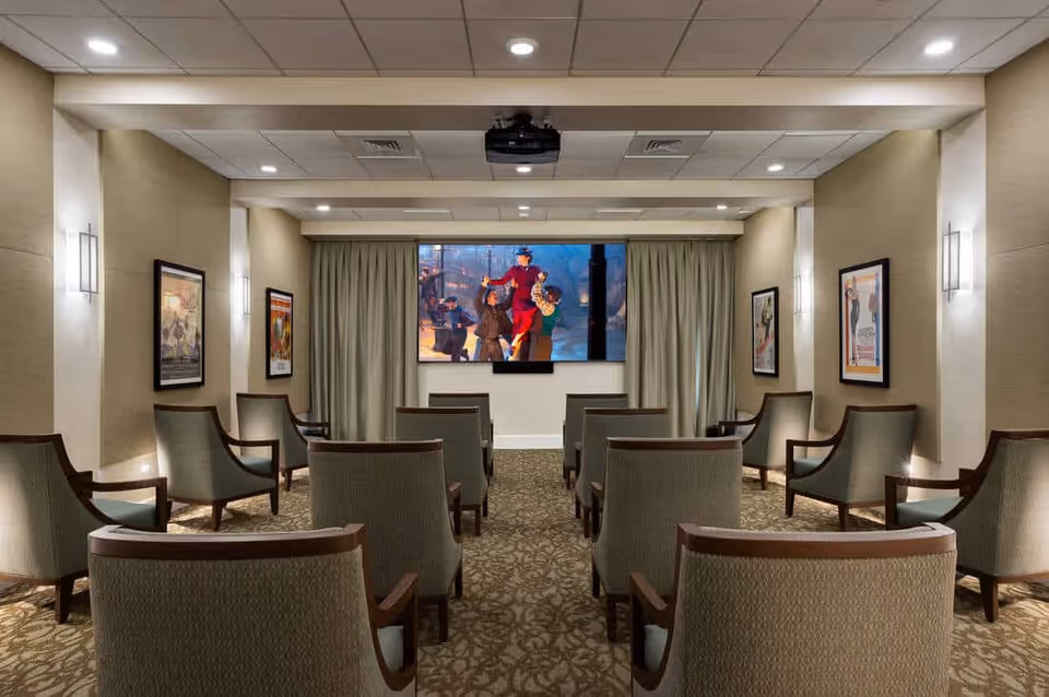 A small theater room with rows of upholstered chairs facing a large screen showing a movie scene. The room has beige walls, framed movie posters on the walls, recessed ceiling lights, and a patterned carpet.