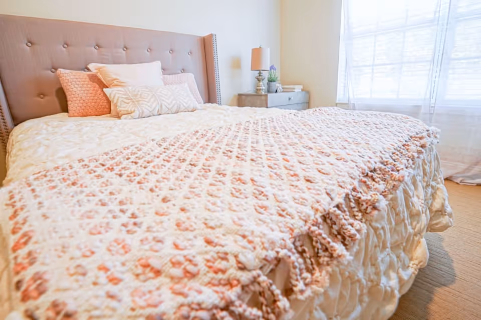 A neatly made bed with a tufted beige headboard, adorned with multiple pillows in shades of white and pink. A textured pink and white blanket is spread across the bed. Next to the bed is a wooden nightstand with a lamp, a small plant, and some books. A window with sheer white curtains allows natural light to brighten the room.