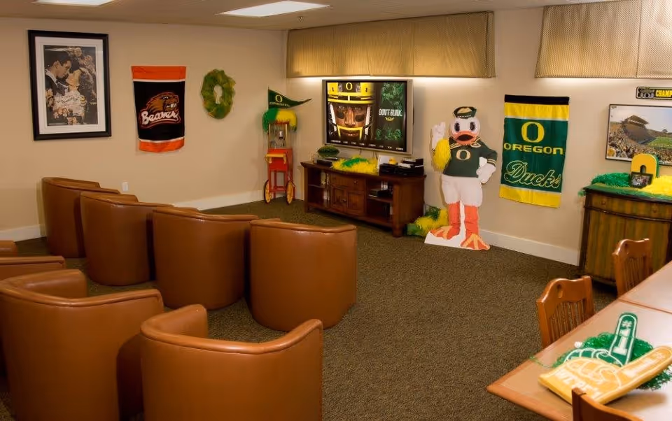 A cozy common area with brown leather chairs arranged in rows facing a TV mounted on the wall. The room is decorated with University of Oregon sports memorabilia, including banners, a cardboard cutout of the mascot, and themed decorations. There is a popcorn machine in the corner and a table with chairs on the right side.