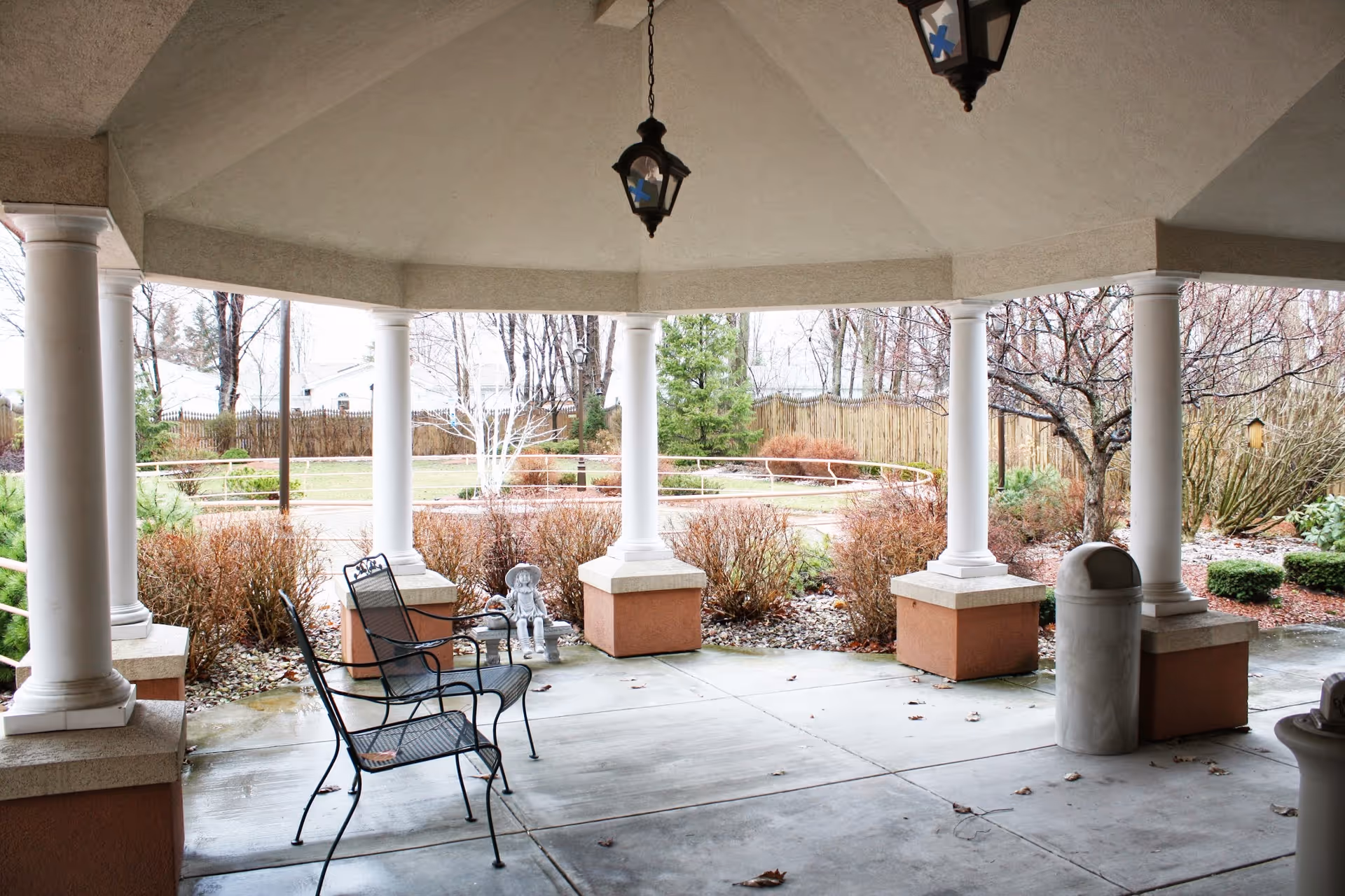 Covered outdoor patio area with white columns and hanging lanterns, two black metal chairs, a small statue, and a trash can. Surrounding the patio are bushes, trees, and a fenced garden area.