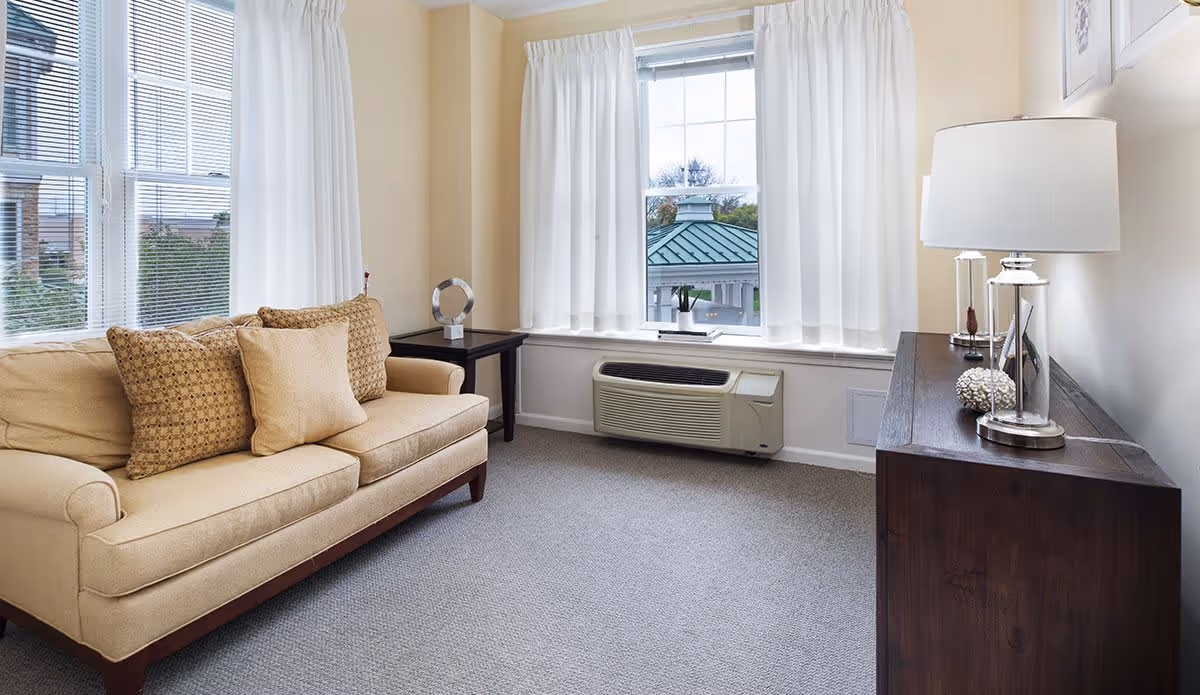 A cozy living room with a beige sofa adorned with three cushions, a dark wooden side table with a decorative item, a large window with white curtains letting in natural light, a wall-mounted air conditioning unit below the window, and a dark wooden cabinet with a modern lamp and decorative objects on top.
