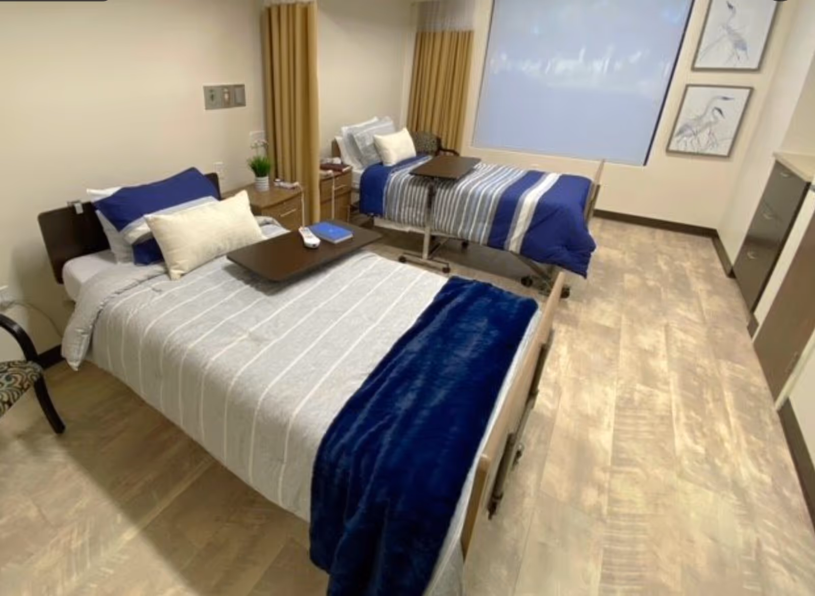 A clean and well-lit room with two single beds, each with a bedside table and an overbed table. The beds have blue and gray bedding with pillows. There is a window with curtains and two framed pictures of birds on the wall. The floor has a light wood pattern, and there is a chair partially visible on the left side.