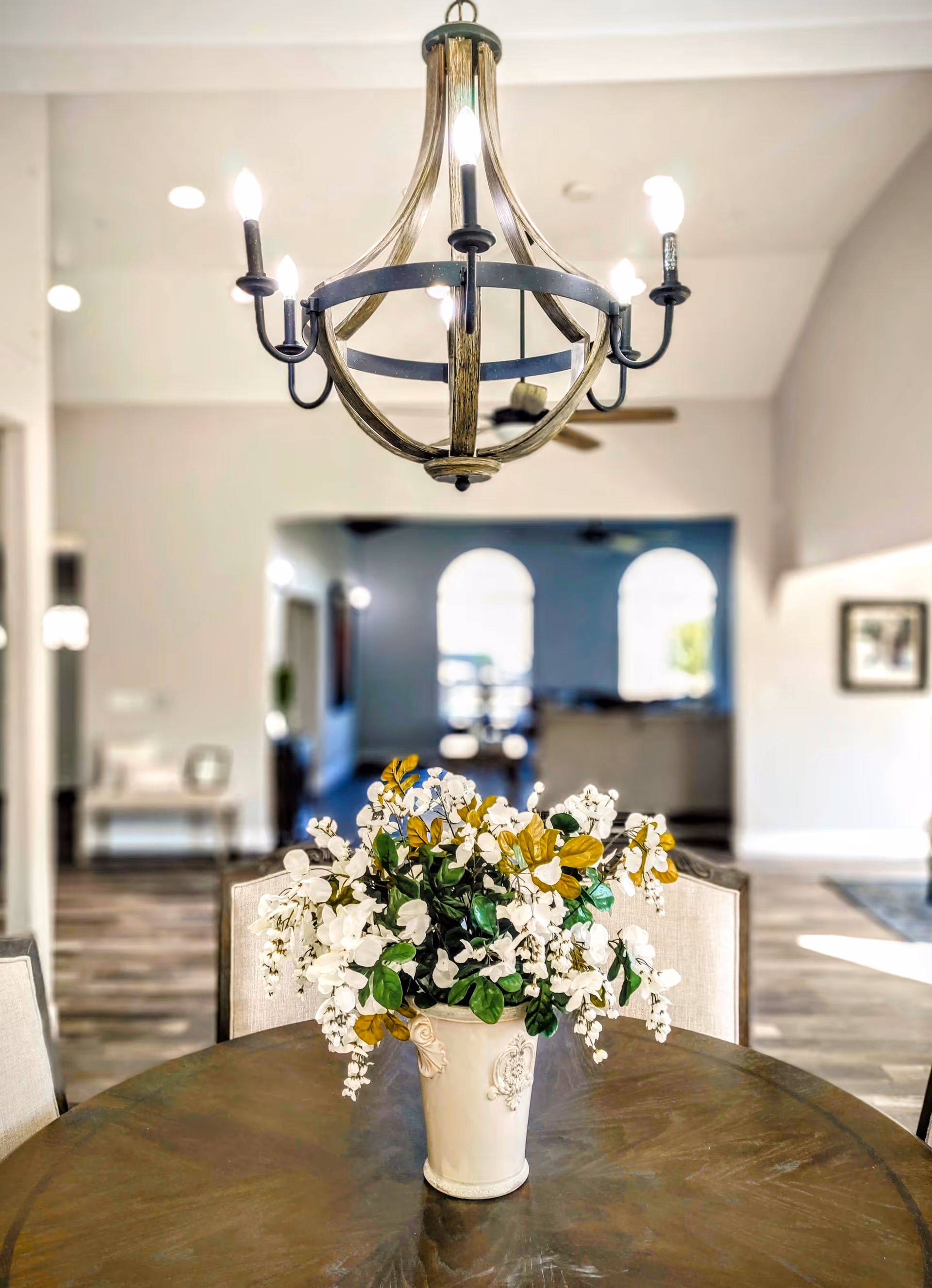 A round wooden table with a white vase filled with white and green flowers in the center. Above the table hangs a rustic chandelier with six candle-like light bulbs. The background shows a spacious, well-lit interior with arched windows and neutral-colored walls.