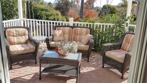 Outdoor patio area with three cushioned wicker chairs and a matching wicker loveseat arranged around a glass-top wicker coffee table with a decorative centerpiece. The patio is enclosed by a white railing and surrounded by greenery and trees under a sunny sky.