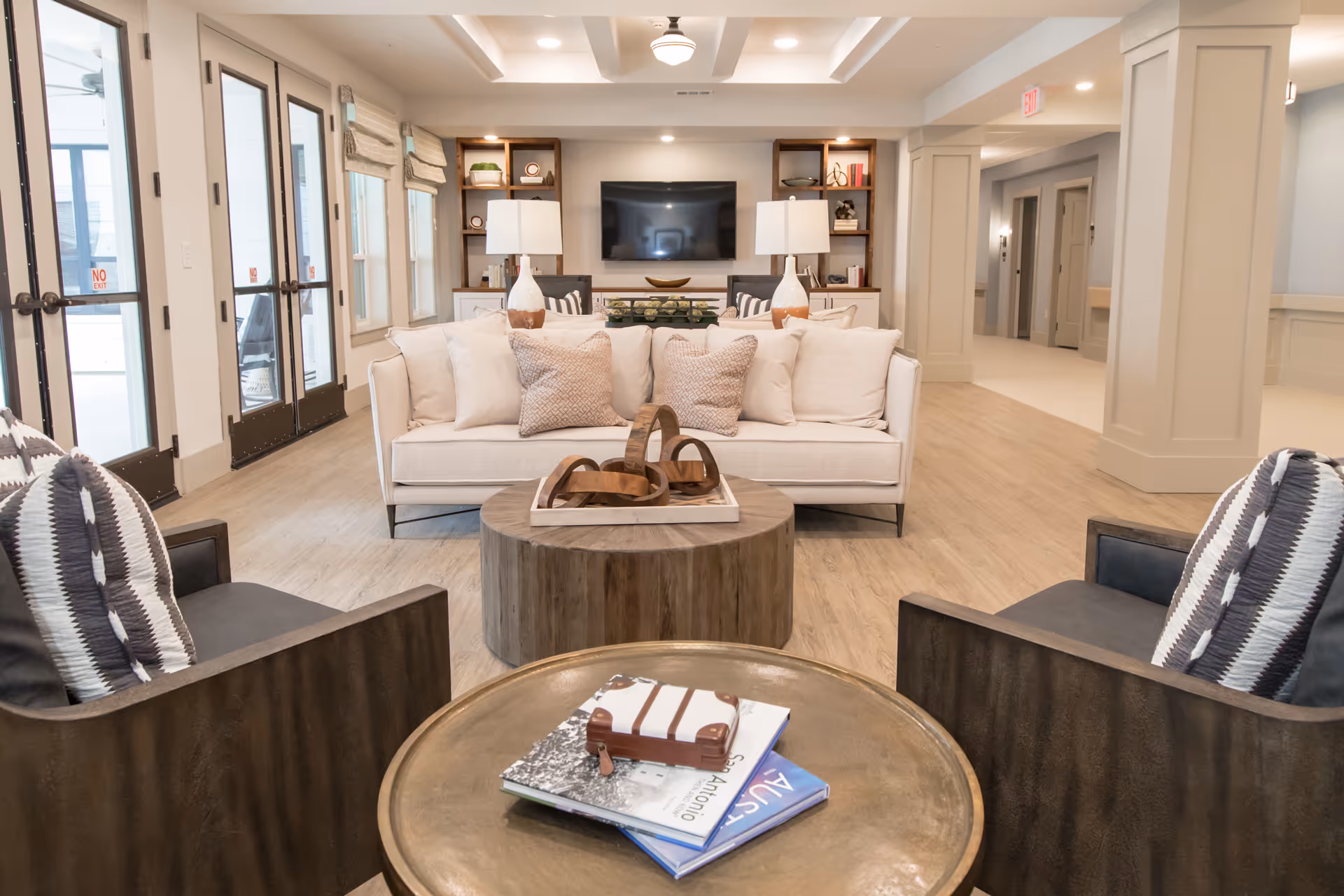 A modern and cozy living room area featuring a white sofa with multiple cushions, two dark armchairs with patterned pillows, a round wooden coffee table with decorative items, and a round side table with books and a small decorative suitcase. The room has light wood flooring, built-in shelves with lamps and decor, a wall-mounted flat-screen TV, and large windows and glass doors letting in natural light.
