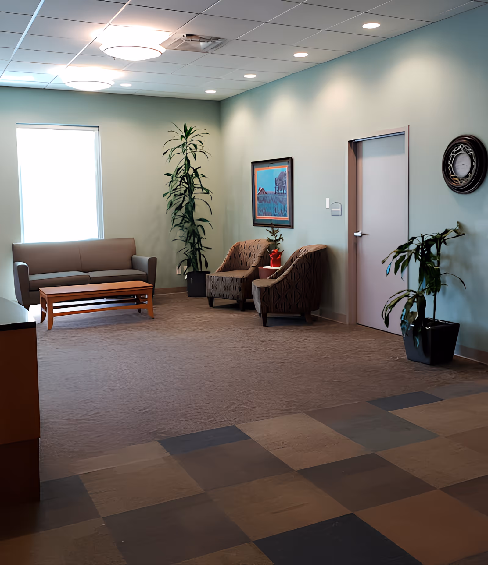 A waiting area with a gray sofa, two patterned armchairs, a wooden coffee table, and several potted plants. The walls are painted light green and decorated with a framed painting and a round wall clock. The floor has a mix of carpet and tiled sections, and the ceiling has recessed lighting and circular light fixtures.