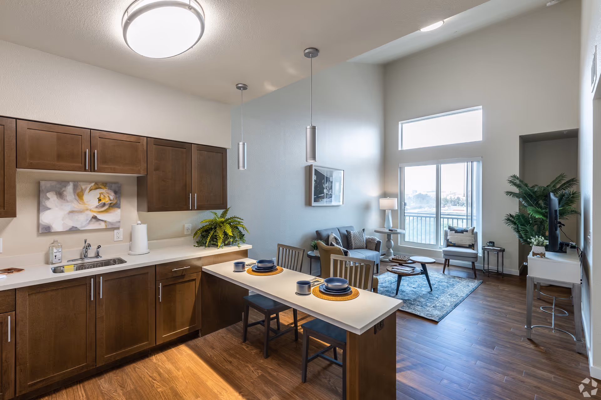 Modern open-concept living space with a kitchen featuring dark wood cabinets, a white countertop island set with blue dishes and cups, and a living room area with a gray sofa, armchair, coffee tables, a floor lamp, and large windows letting in natural light.