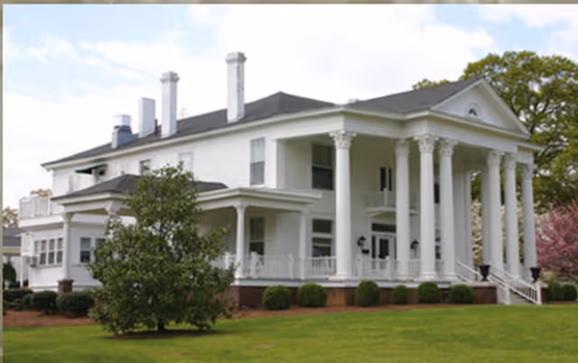 A large white two-story building with classical architectural features including tall columns and a triangular pediment, surrounded by green grass and trees under a partly cloudy sky.
