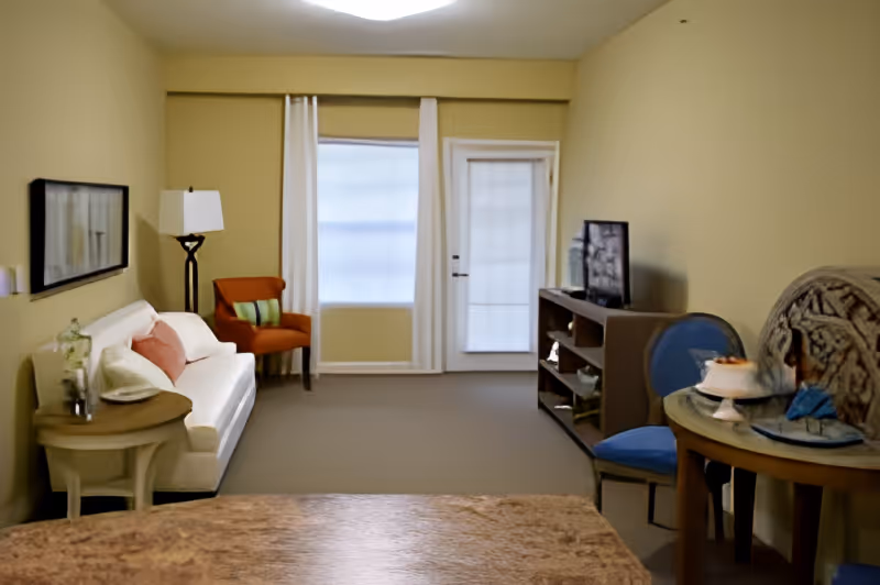 Furnished living area with a sofa and side table on the left, an armchair by a window and a TV console with a small round table and chairs on the right.