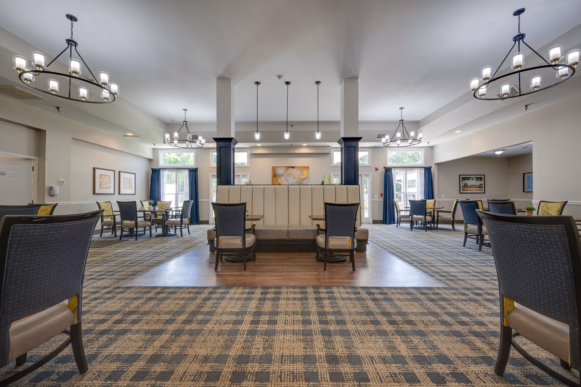 Spacious and well-lit dining area in an assisted living facility with multiple tables and chairs arranged neatly. The room features patterned carpet flooring, modern chandeliers, large windows with blue curtains allowing natural light, and a central seating area with a cushioned bench. Artwork is displayed on the walls, and the overall ambiance is welcoming and comfortable.
