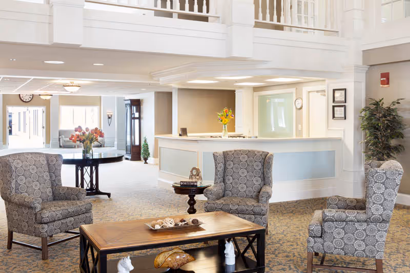 A bright and spacious senior living facility common area with three patterned armchairs arranged around a wooden coffee table. The room features a reception desk with a vase of flowers, a tall grandfather clock, and a round table with another vase of flowers. The area has light-colored walls, carpeted floors, and a second-floor balcony overlooking the space.
