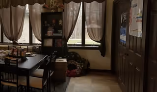 Small dining area with a table and chairs by large curtained windows, a shelving unit with items, and a closed interior door.