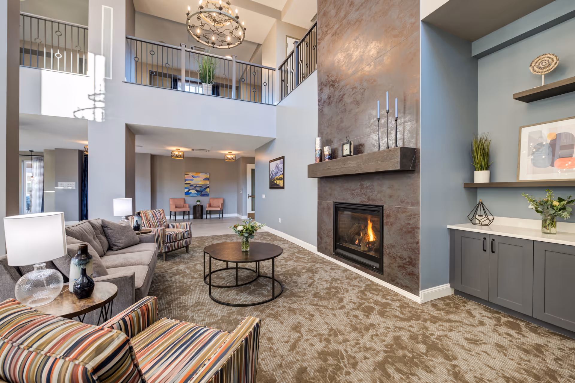 A spacious and well-lit living room area with a high ceiling and a balcony railing on the upper level. The room features a modern fireplace with a dark stone surround and a wooden mantel decorated with candles and a clock. There is a round coffee table with a flower vase in the center, surrounded by a gray sofa and two striped armchairs. The carpet has a patterned design, and the walls are painted in soft gray and blue tones. Built-in cabinets and shelves with decorative items and plants are visible on the right side.