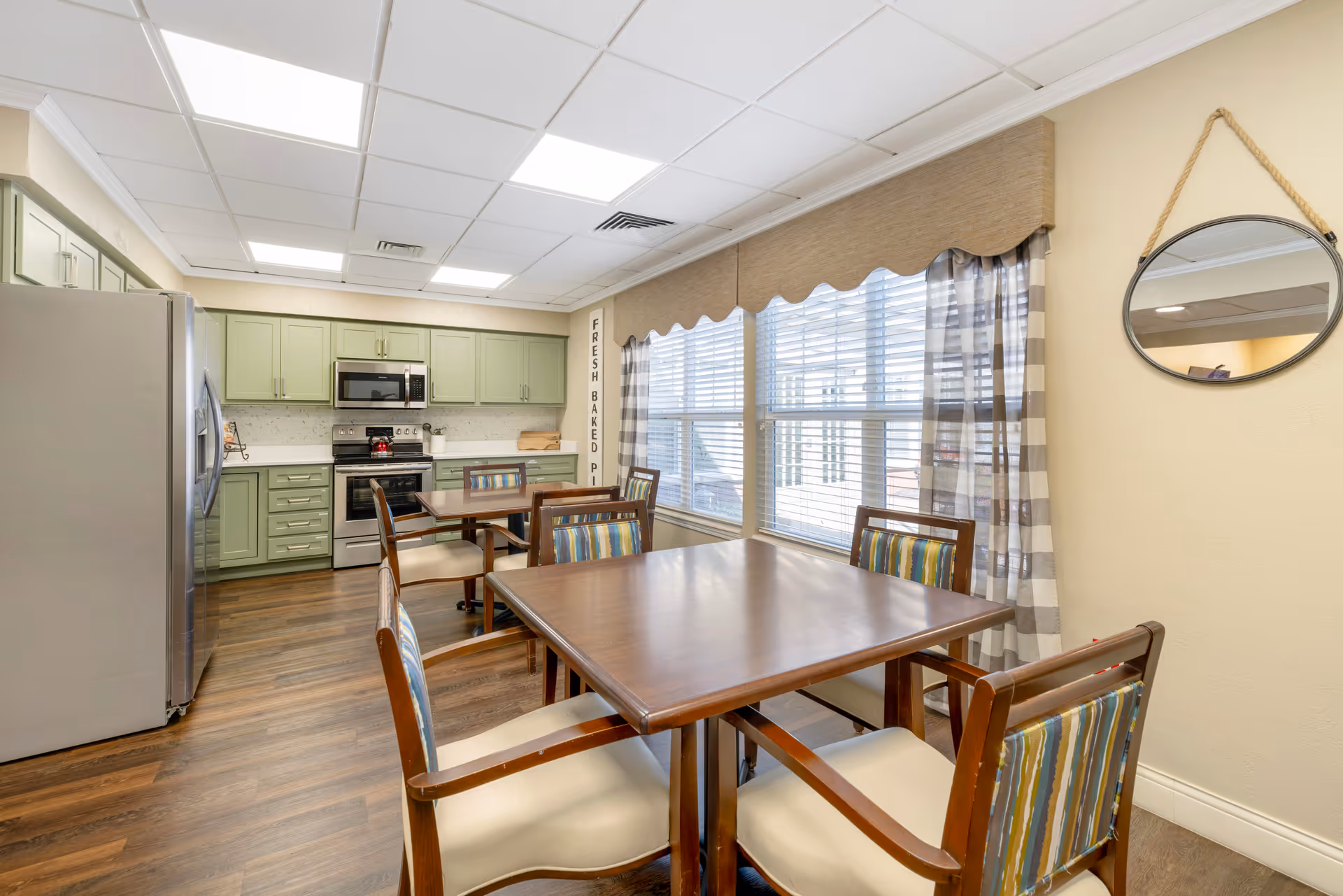 Communal dining room with wooden tables and upholstered chairs, large windows with curtains, and a kitchen with green cabinets and stainless appliances.