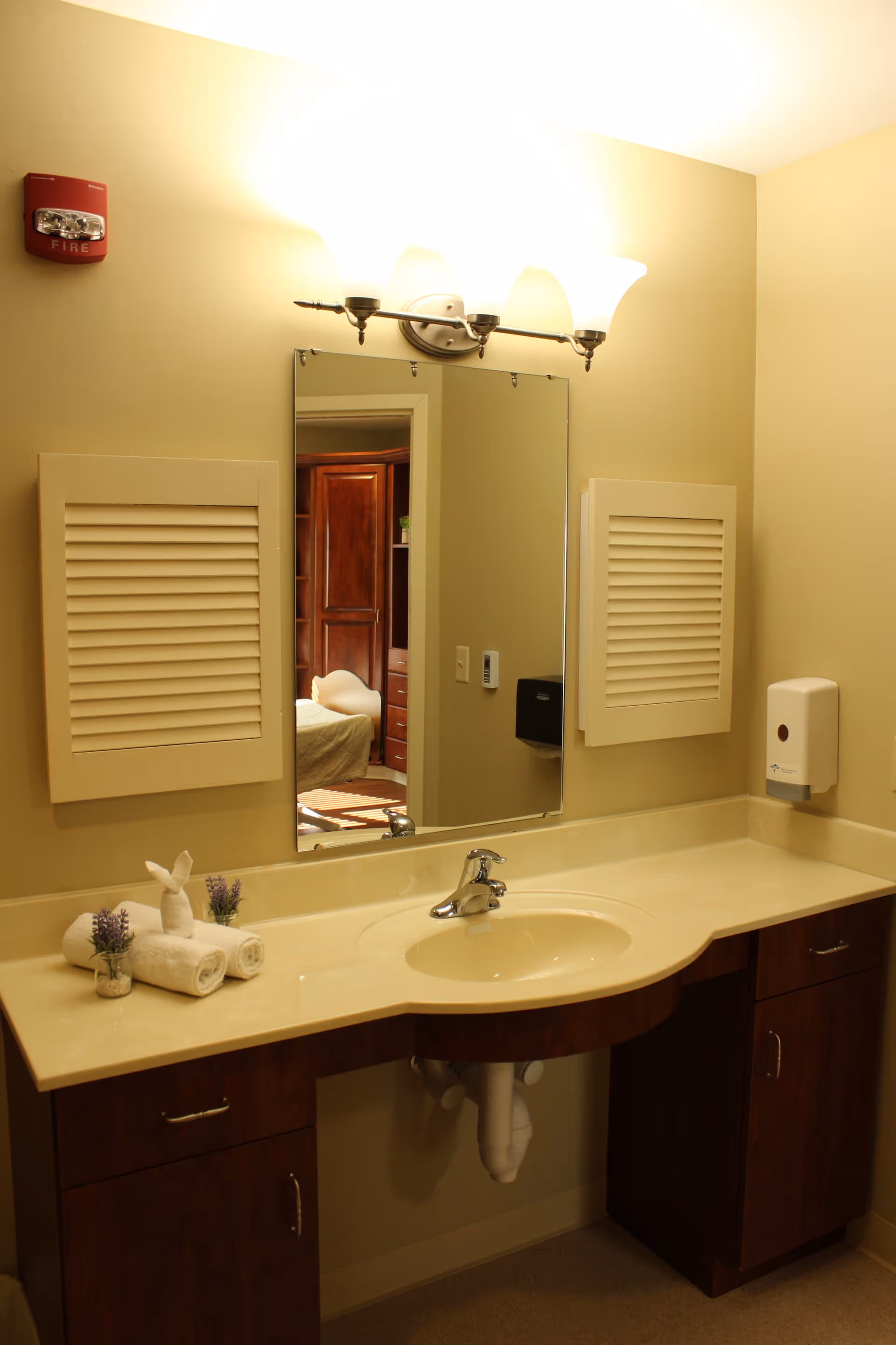 A bathroom vanity with a sink, faucet, and countertop. Above the sink is a rectangular mirror with a three-light fixture mounted above it. On the countertop, there are rolled white towels and small decorative plants. The walls are beige, and there are two white vent covers on either side of the mirror. A soap dispenser is mounted on the wall to the right of the sink. The reflection in the mirror shows part of a bedroom with wooden furniture.