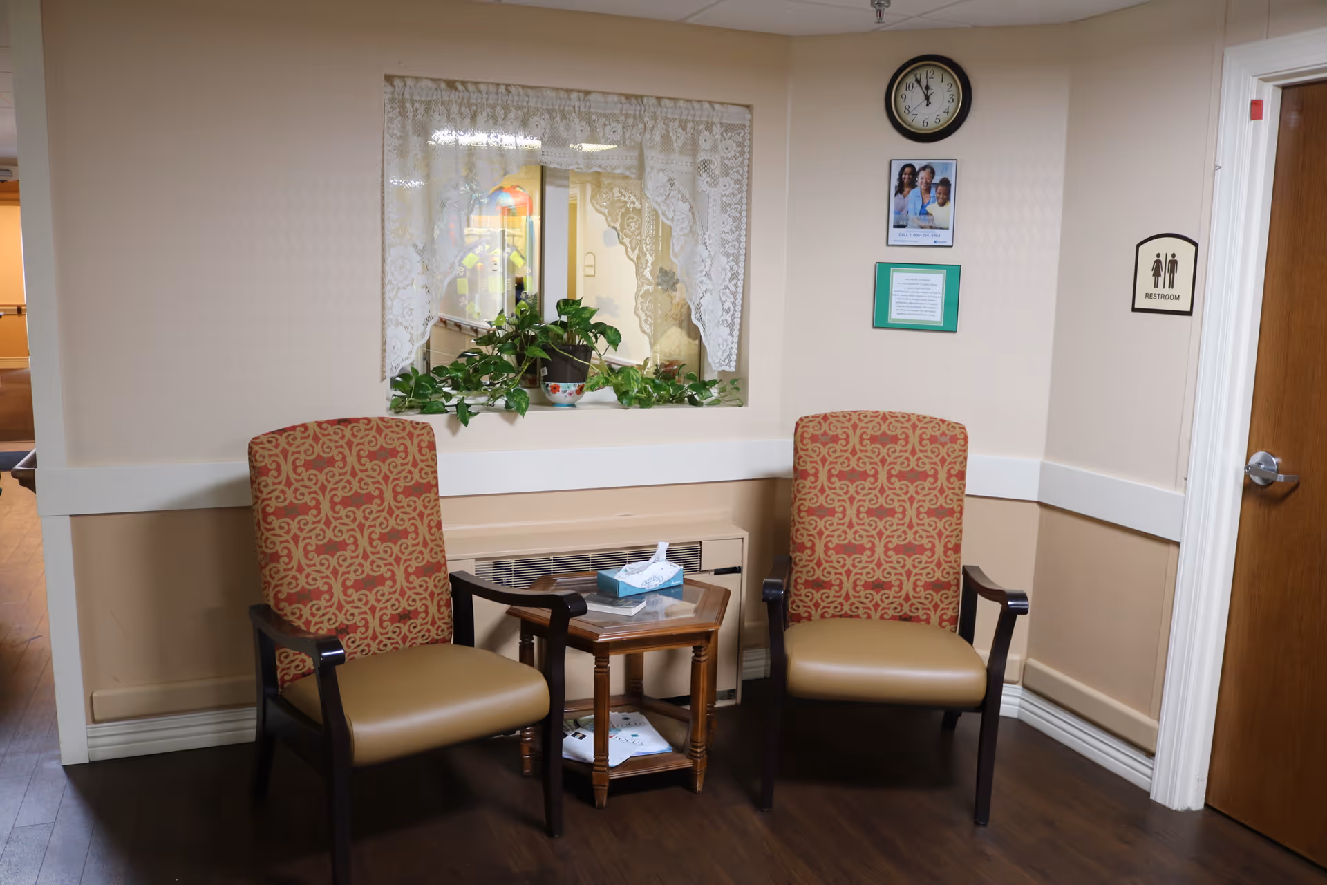 Two patterned armchairs and a small side table in a senior living seating nook with a potted plant, wall clock, and restroom sign.