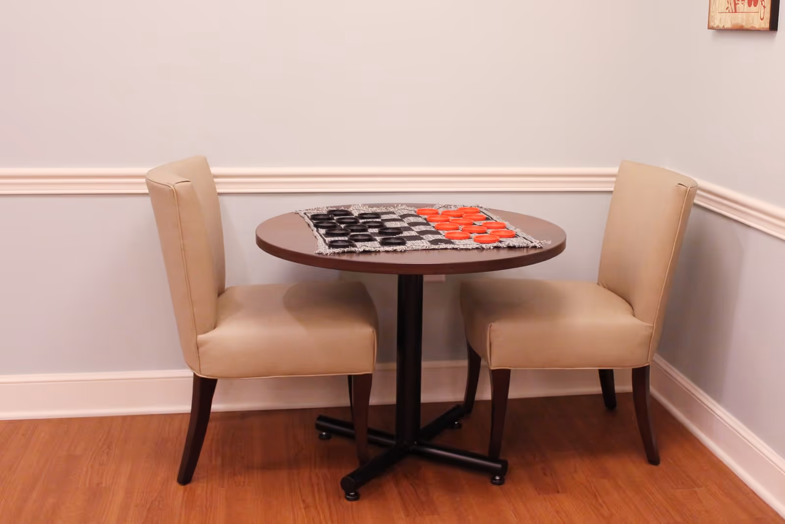 A small round wooden table with a checkers board and red and black checkers pieces set up for a game, flanked by two beige upholstered chairs, against a light blue wall with white trim and wooden floor.