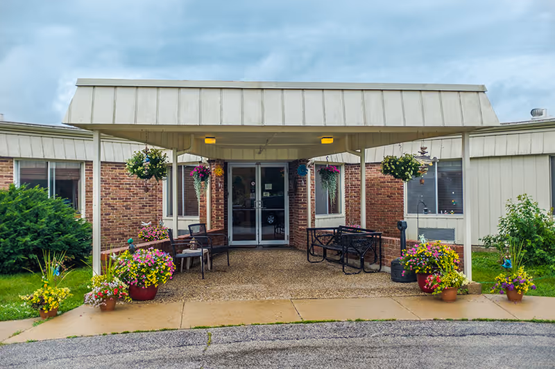 Entrance to a building with a covered porch area featuring hanging flower baskets and potted plants. There are chairs and benches under the porch, with brick walls and glass double doors leading inside. The sky is cloudy.