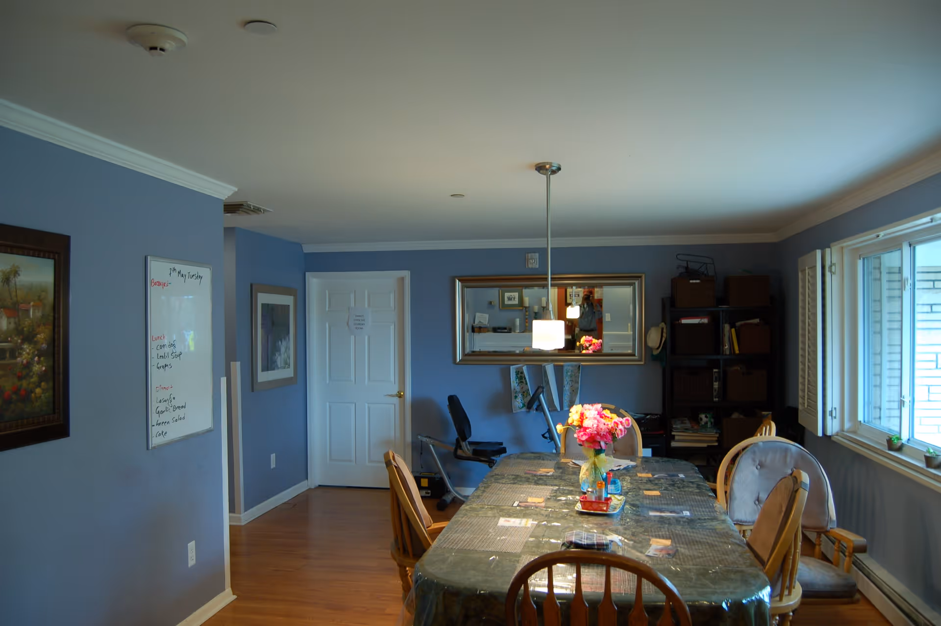 A dining room with a long table covered with a plastic tablecloth and a vase of pink flowers in the center. There are several wooden chairs around the table. The walls are painted blue and decorated with framed pictures and a whiteboard. A large mirror hangs on the far wall above an exercise bike. A window on the right side lets in natural light, and there is a shelving unit with baskets and books in the corner.