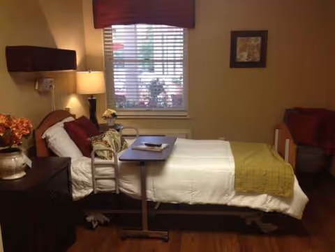 Single patient bedroom with bed, overbed table, chairs, dresser, and a window with blinds.