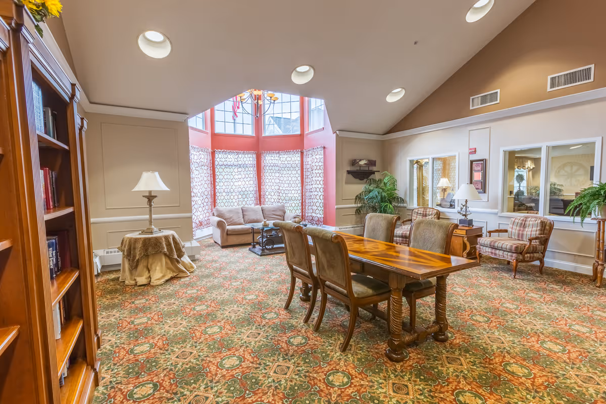 A spacious and well-lit living room area with a large wooden table and six upholstered chairs in the center. The room features patterned carpet, a bookshelf filled with books on the left, a small round table with a lamp, and a seating area with a beige sofa and two plaid armchairs near large windows with patterned curtains. The ceiling has recessed lighting and a chandelier, and there are plants and decorative items around the room.