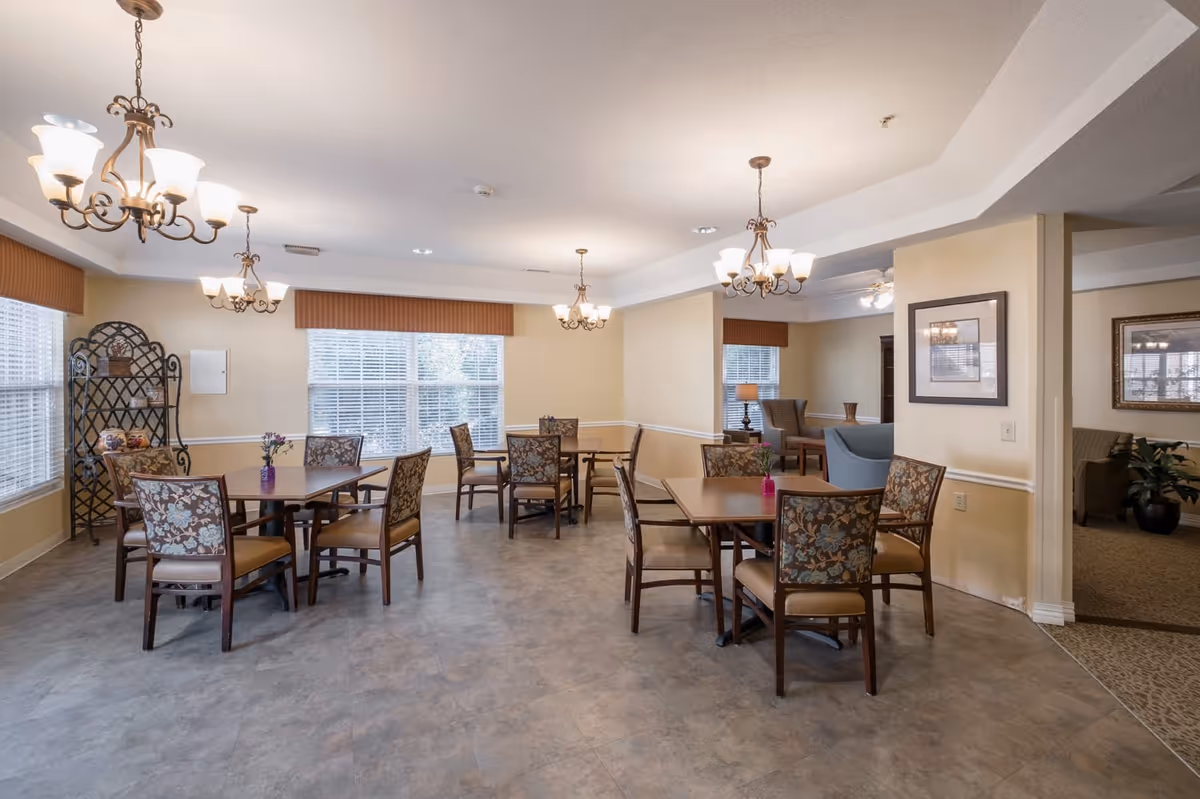 A well-lit dining area in an assisted living facility with several wooden tables and floral upholstered chairs. The room has large windows with blinds, decorative light fixtures hanging from the ceiling, and small vases with flowers on the tables. In the background, there is a seating area with armchairs and framed artwork on the walls.