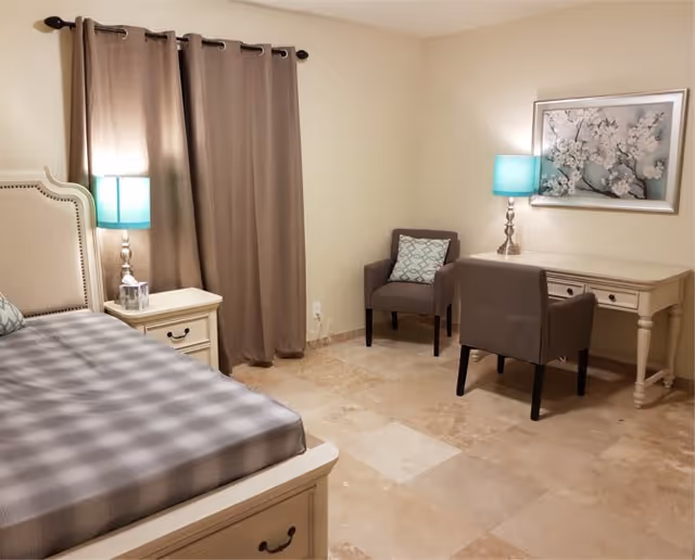 A neatly arranged bedroom with a bed on the left side, a nightstand with a lamp next to it, beige curtains covering a window, two chairs with a cushion, a desk with a lamp, and a framed floral artwork on the wall.