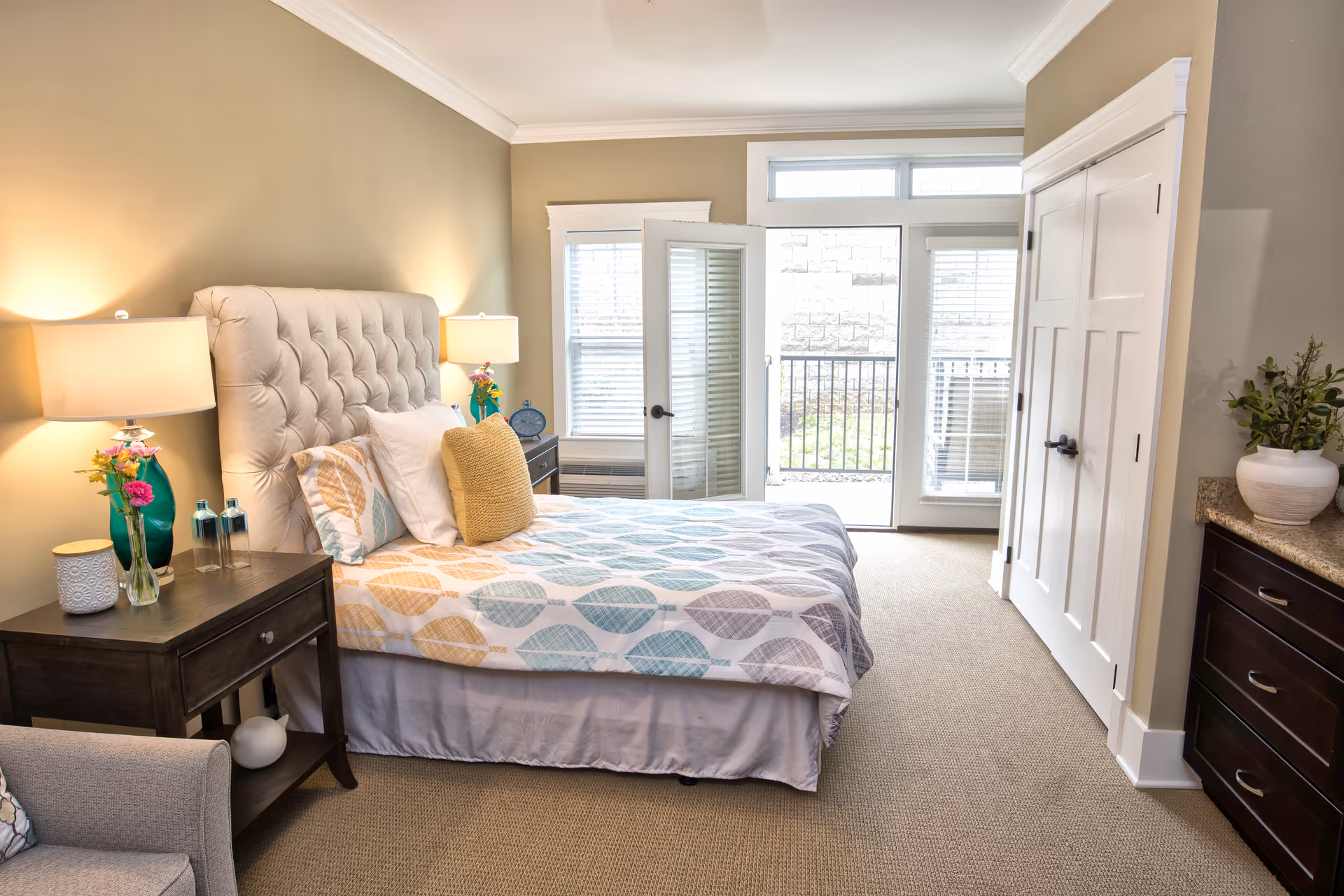 A bright and cozy bedroom with a large bed featuring a tufted headboard and patterned bedding. There are two bedside tables with lamps and decorative items, a comfortable armchair, a closet with double doors, and a dresser with a plant on top. The room has beige walls, carpeted floor, and a door leading to an outdoor area with a railing.