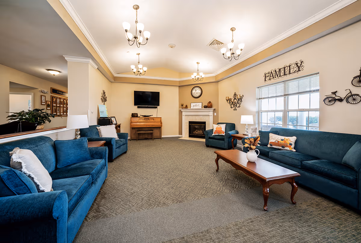 A spacious living room with blue upholstered sofas and armchairs arranged around a wooden coffee table. The room features a fireplace with a clock above it, a wall-mounted TV, and a piano. There are decorative wall hangings including a metal 'FAMILY' sign above a large window, and two chandeliers hanging from the ceiling. The room has beige walls and carpeted flooring.