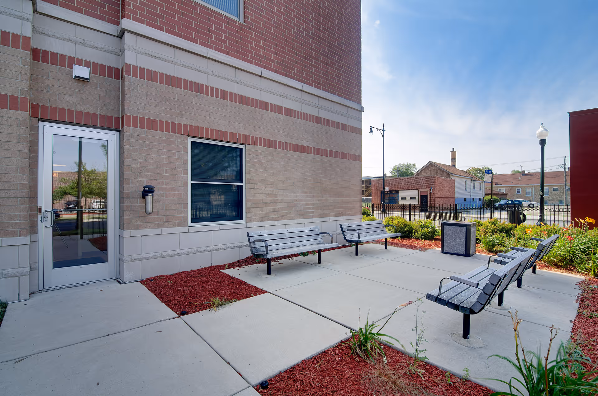 Outdoor seating area at Senior Suites of Auburn Gresham with four wooden benches arranged on a concrete patio next to a brick building. There are flower beds with red mulch and some plants surrounding the patio. A trash bin and street lamps are visible in the background under a clear blue sky.