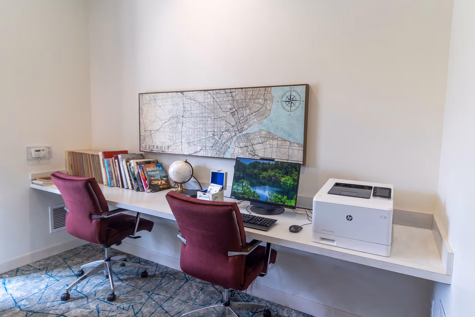 A small office or study area with a long white desk mounted to the wall. Two maroon swivel chairs are positioned in front of the desk. On the desk, there is a computer monitor, keyboard, mouse, a white HP printer, a globe, and several books. Above the desk, a framed map of Detroit is hung on the wall. The floor is carpeted with a blue and gray patterned carpet.