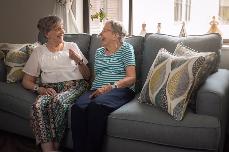 Two elderly women sit and laugh together on a couch in a cozy living room.