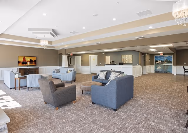 A spacious and well-lit living room area in a senior living facility with multiple seating arrangements including sofas and armchairs. The room features a carpeted floor, neutral-colored walls, recessed lighting, a chandelier, and a flat-screen TV mounted on the wall. There is a reception desk area in the background and a large mural depicting a waterfront scene.