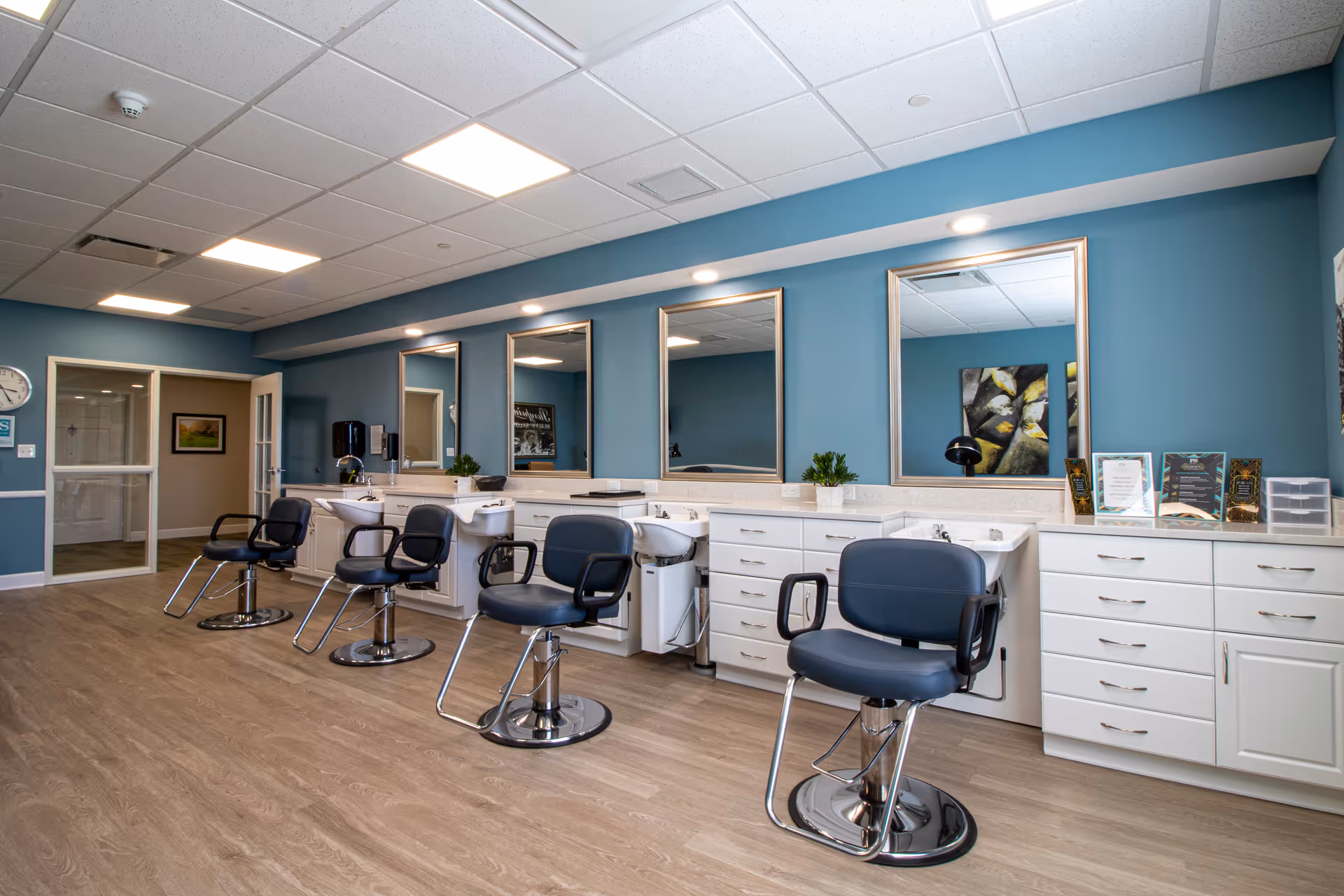 Interior view of a salon area with four black salon chairs in front of white counters with sinks and large mirrors mounted on a blue wall. The floor is wood-style, and the ceiling has recessed lighting panels. There are small plants and framed certificates on the counter.
