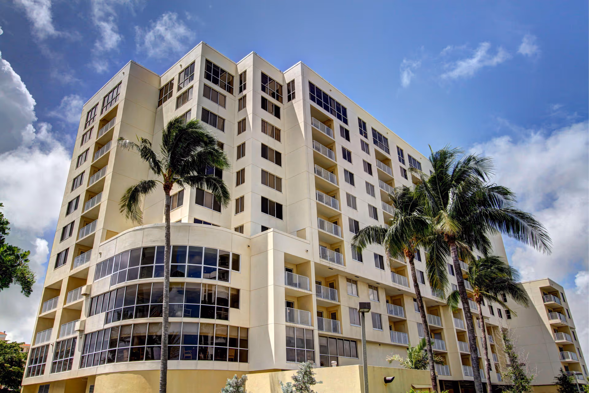 Exterior view of a multi-story residential building with balconies and large windows, surrounded by palm trees under a partly cloudy blue sky.