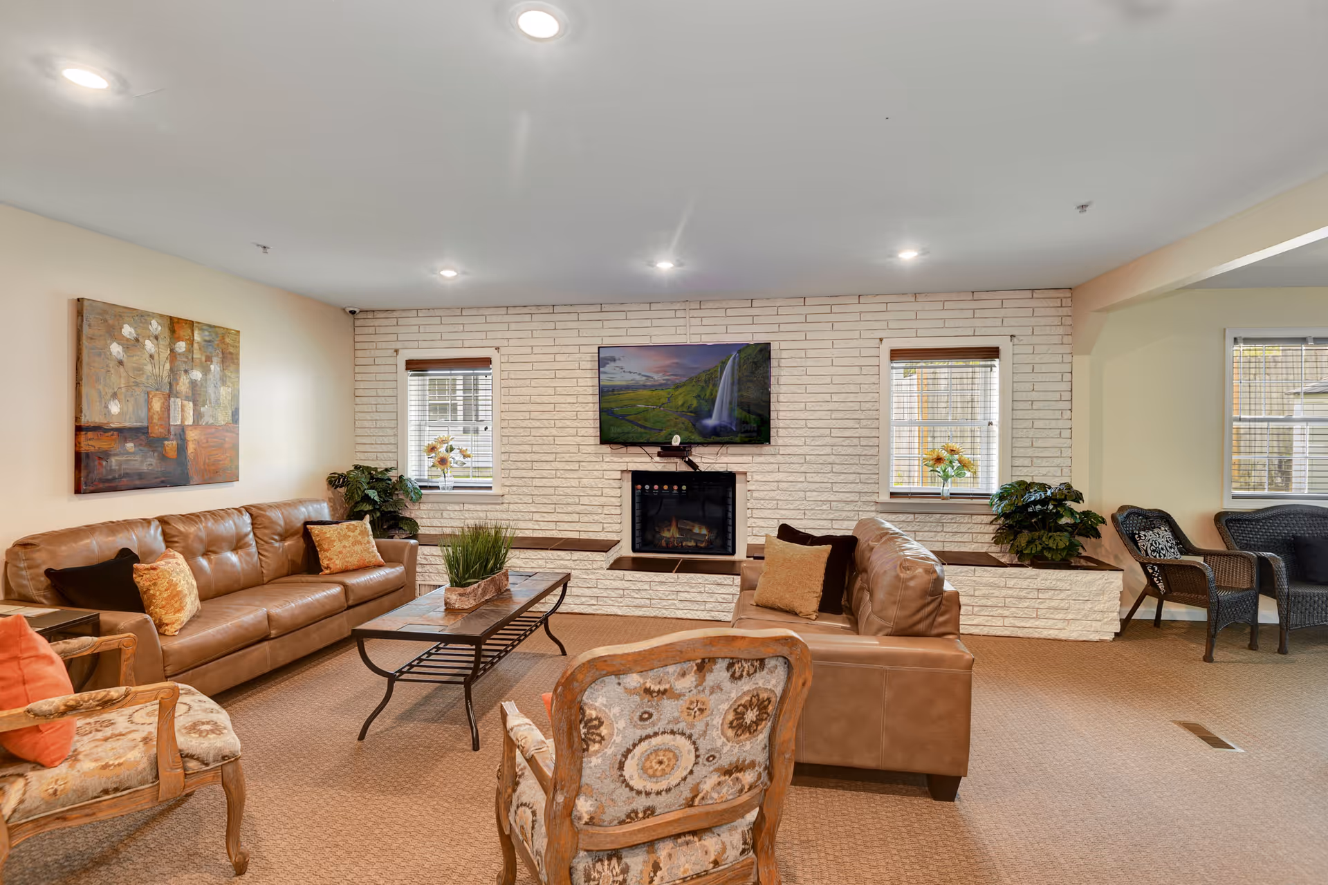 A cozy living room with two brown leather sofas, two patterned armchairs, a rectangular coffee table with a plant centerpiece, and a wall-mounted TV above a fireplace set in a white brick wall. There are two windows with blinds and flower vases on the windowsills, along with some green plants. The room is warmly lit with recessed ceiling lights and has beige carpet flooring.