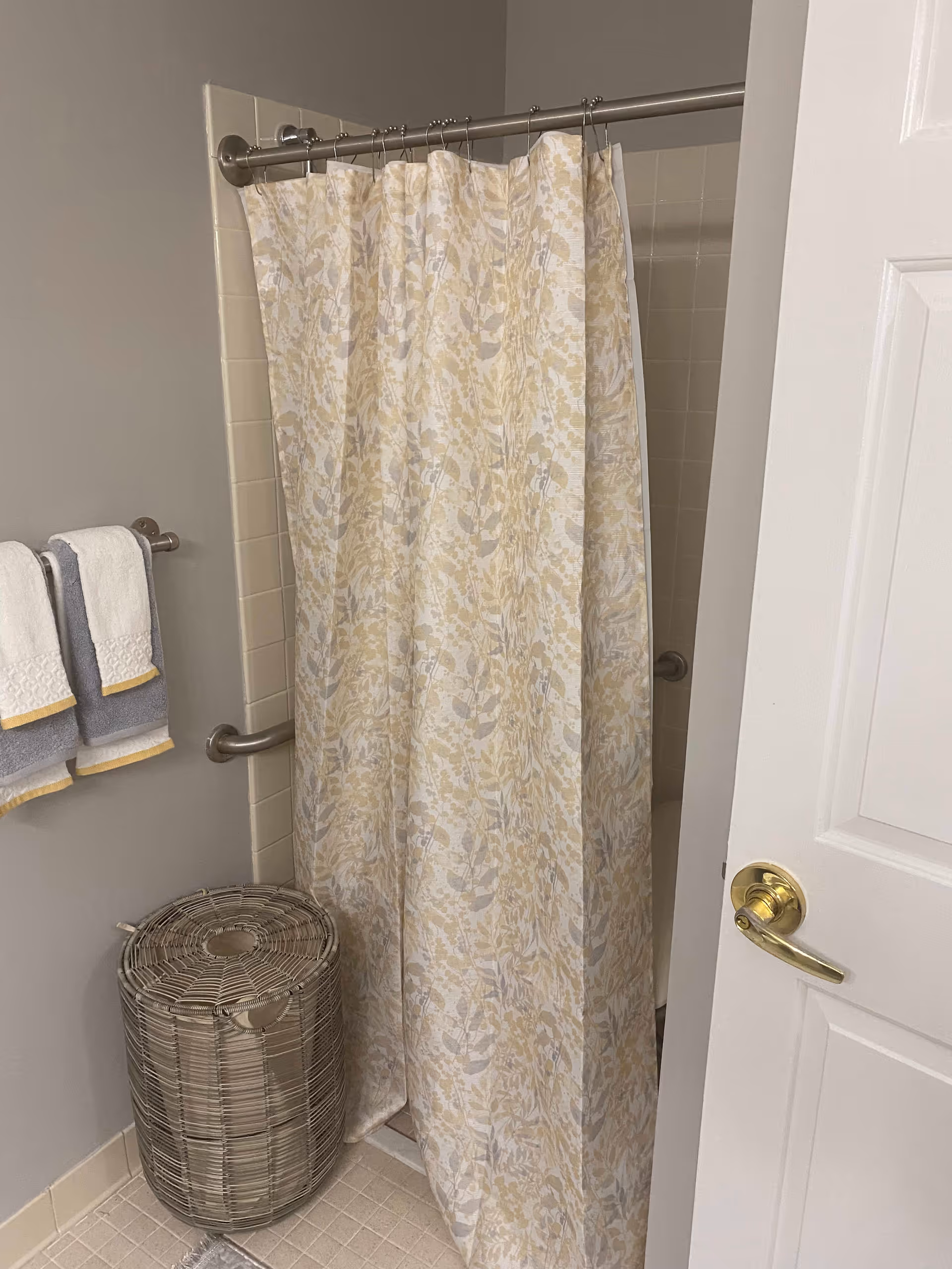 Bathroom with a shower area covered by a floral-patterned shower curtain in beige and gray tones. There are two towel racks on the left wall holding white and gray towels with yellow trim. A woven laundry basket is placed on the floor next to the shower. The bathroom door with a gold-colored handle is partially open on the right side.