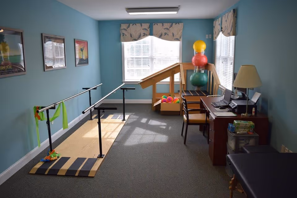 A therapy or rehabilitation room with parallel bars for walking exercises, colorful therapy balls stacked in a corner, a small wooden ramp, a desk with a computer and lamp, and three framed motivational posters on a blue wall.