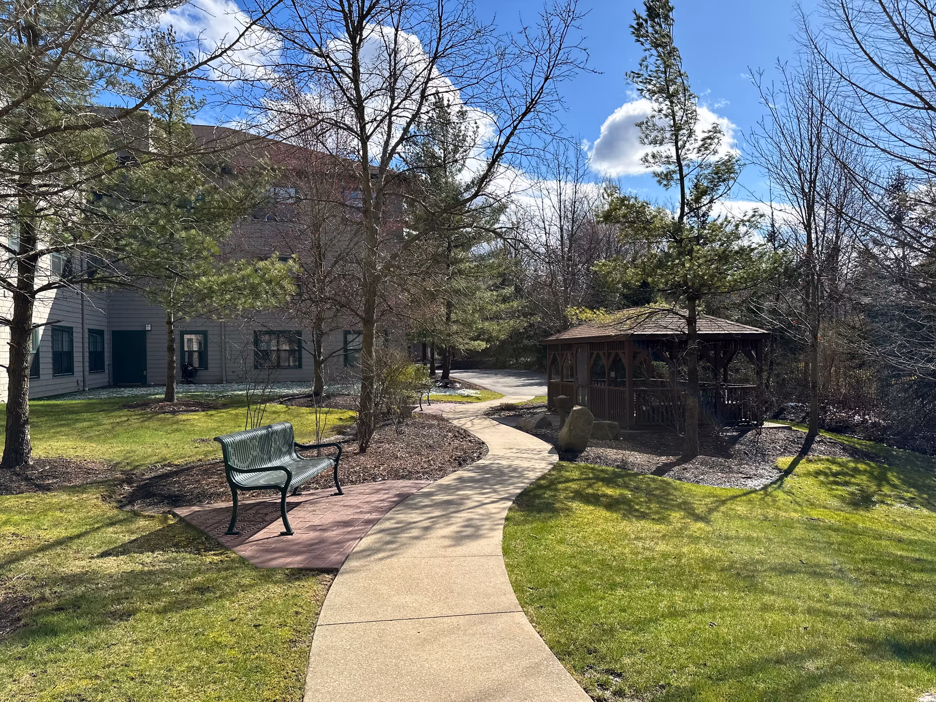 Curved walkway leading past a bench to a wooden gazebo in a landscaped courtyard beside a multi-story building under a blue sky.