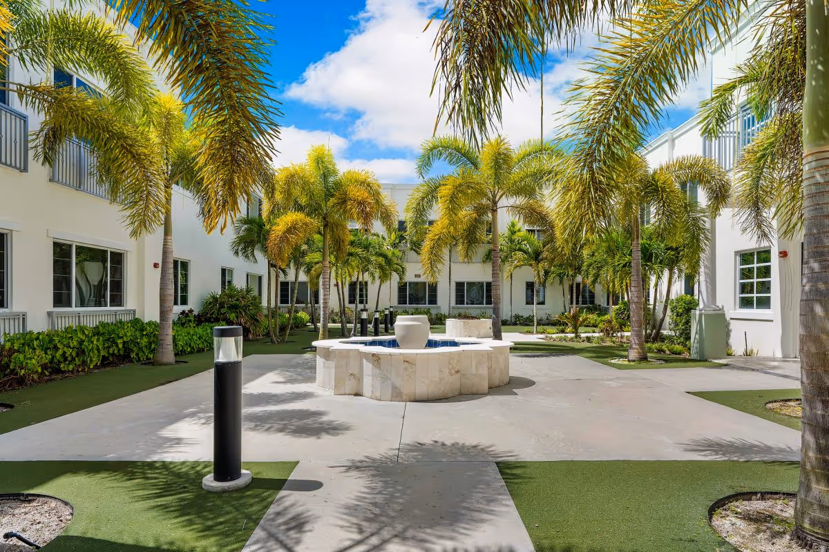Outdoor courtyard area at Luxe Senior Living at Jupiter featuring a central stone fountain surrounded by palm trees, green landscaping, and white buildings under a partly cloudy blue sky.