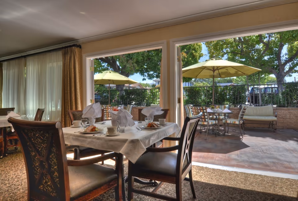 A dining area inside a memory care community with a table set for four, including plates with food, cups, and folded napkins. Large open sliding doors lead to an outdoor patio with tables, chairs, and umbrellas surrounded by greenery and trees.