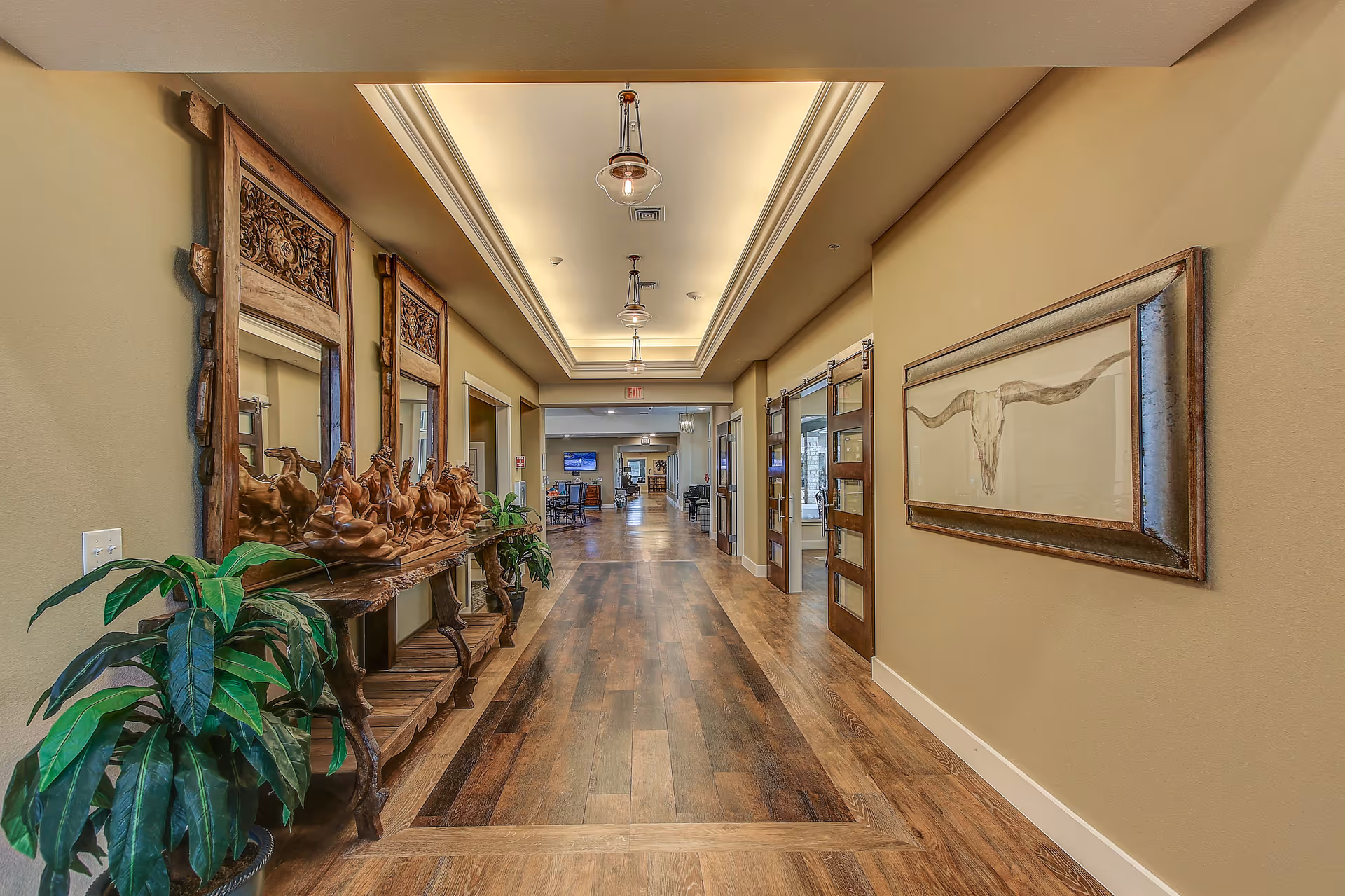 A long hallway in a senior living facility with wooden flooring and beige walls. On the left side, there is a decorative wooden console table with a sculpture of running horses and two large mirrors above it. There are also two green potted plants near the table. On the right wall, there is a framed artwork of a longhorn skull. The hallway is well-lit with ceiling lights and leads to a common area with chairs and a TV in the distance.