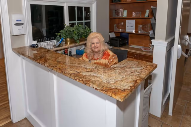 A woman with blonde hair sitting behind a granite reception desk in an indoor office area with wooden shelves and a window with plants on the counter.