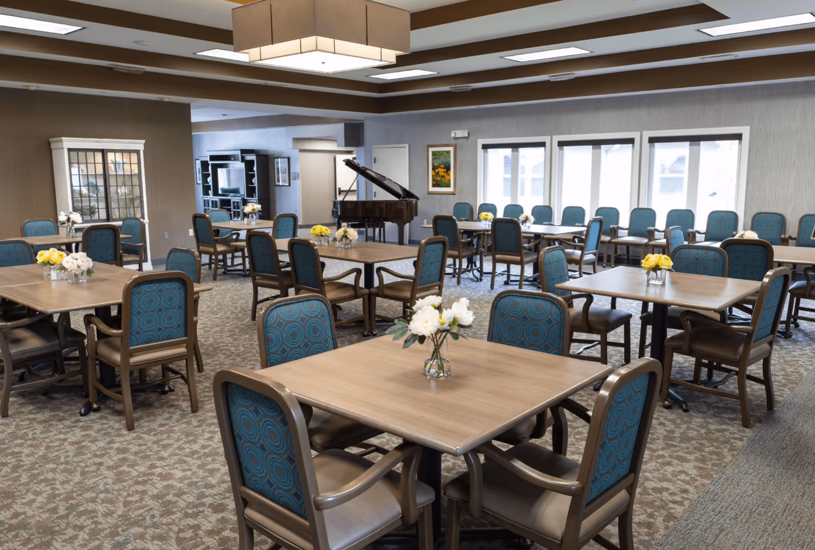 A spacious dining room with multiple square wooden tables surrounded by blue cushioned chairs. Each table has a small vase with white or yellow flowers. The room features large windows letting in natural light, a grand piano in the background, and a cabinet with glass doors on the left side. The ceiling has recessed lighting and a large rectangular light fixture.
