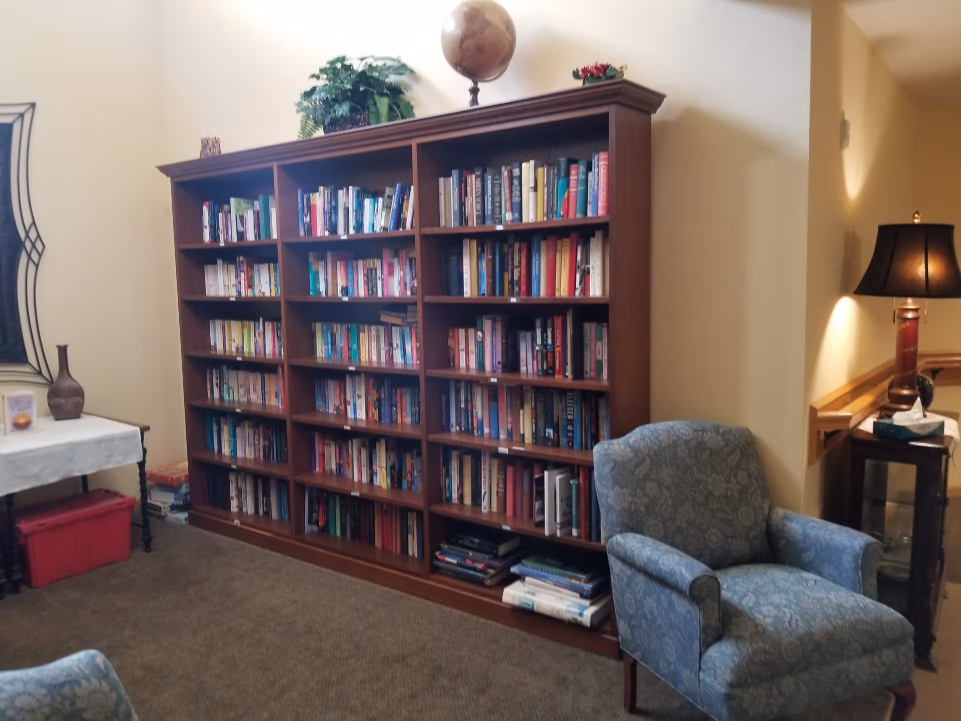 Cozy interior sitting area with a large wooden bookshelf filled with books, an upholstered armchair, and a side table with a lamp.
