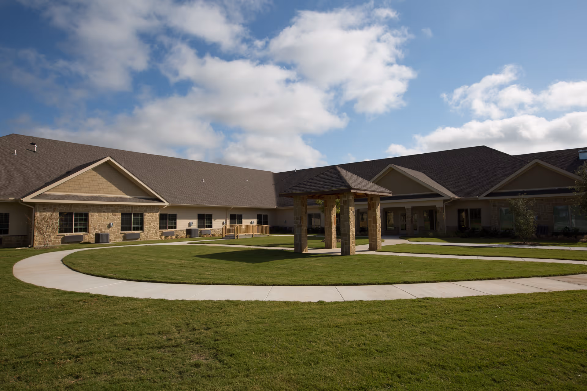 Outdoor courtyard area of Legend Oaks Healthcare and Rehabilitation - Waxahachie featuring a circular concrete walkway surrounding a grassy lawn with a small pavilion structure in the center, and the facility building in the background under a partly cloudy sky.