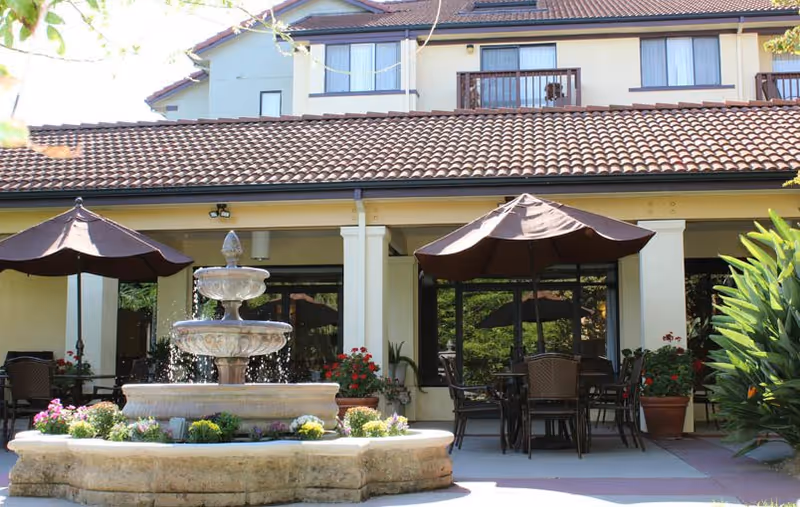 Outdoor patio area at Drake Terrace featuring a multi-tiered stone water fountain surrounded by flowers, several tables with chairs under large brown umbrellas, and a building with a tiled roof and balconies in the background.
