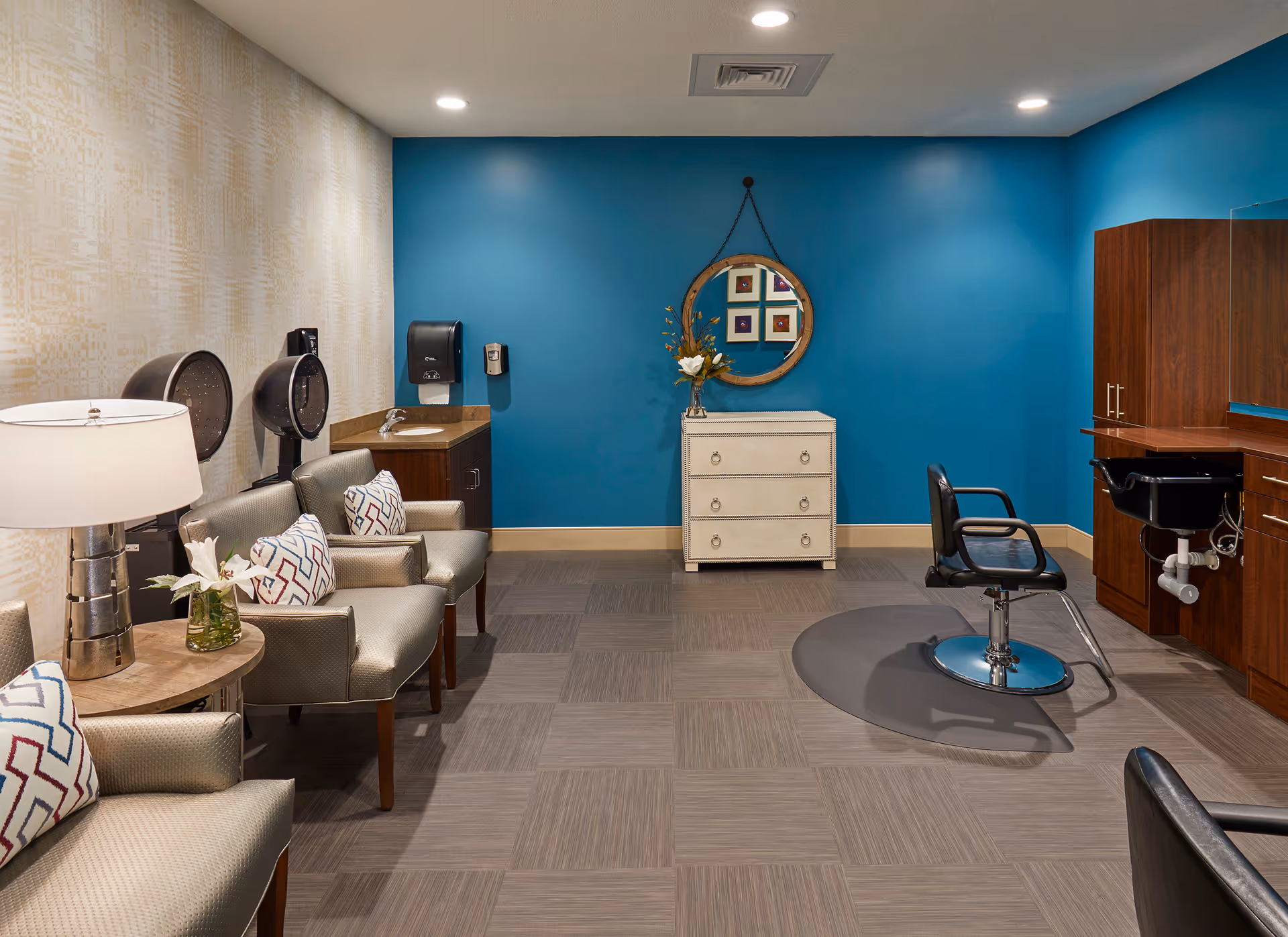 Interior of a salon room with a blue accent wall, a round mirror hanging above a white chest of drawers with a flower vase on top, a black salon chair on a protective mat in front of a wooden counter with a sink, and a seating area with three beige armchairs and a side table with a lamp and flower vase.