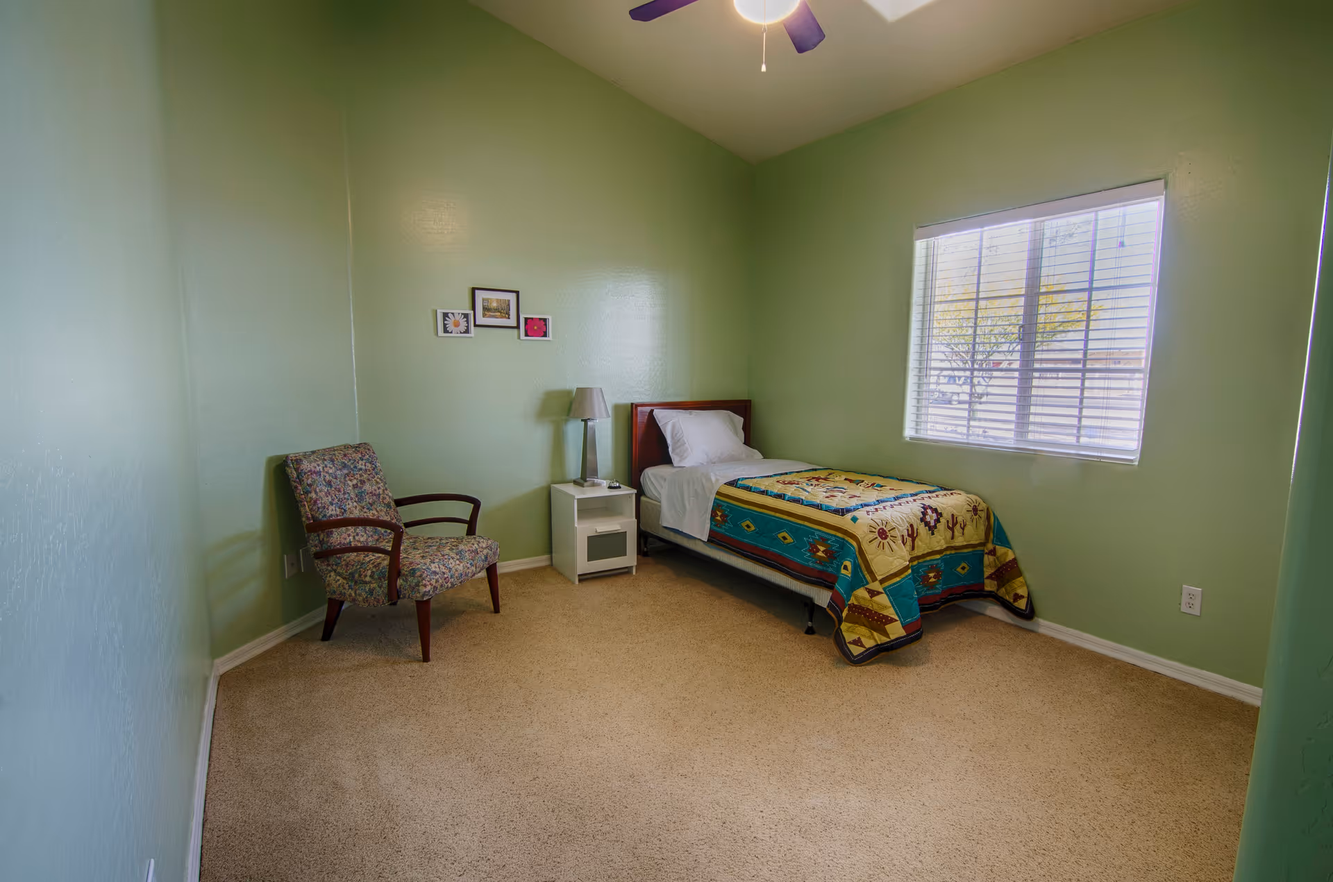 A small bedroom with light green walls and beige carpet. The room contains a single bed with a colorful quilt, a white nightstand with a lamp, a floral upholstered armchair, and three small framed pictures on the wall. A window with white blinds lets in natural light.
