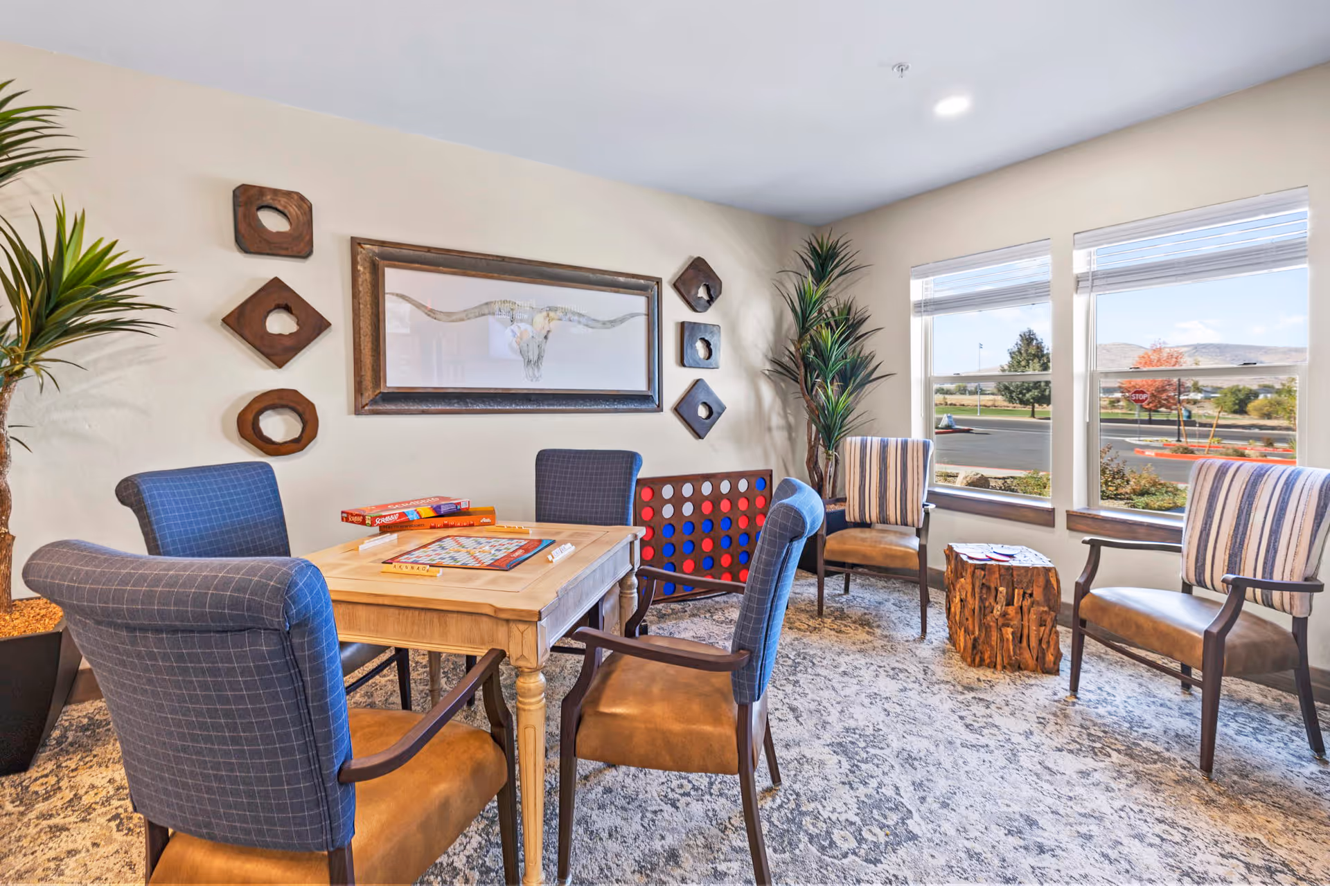 A bright and cozy common area with a wooden table surrounded by four upholstered chairs. On the table are board games including Scrabble and a large Connect Four game. The room has large windows letting in natural light, a patterned carpet, potted plants, and wall decorations including a framed longhorn skull and wooden geometric shapes.